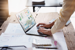 Person analysing financial charts on a laptop with printed graphs, calculator, and documents spread across a wooden desk in a well-lit office.