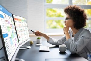 A focused individual in glasses works at a desk, pointing at dual monitors displaying calendars and charts, conveying professionalism and concentration.
