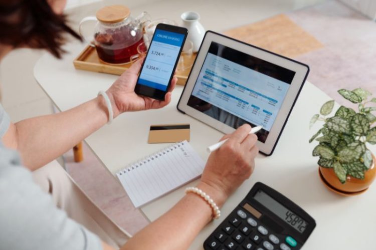 A person sits at a table managing finances. They hold a phone displaying online banking and a tablet with financial statements. Nearby are a notebook, calculator, plant, and teapot.