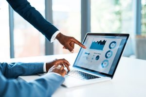Two people in business attire collaborate, with one pointing at data visualizations on a laptop screen while the other operates the keyboard