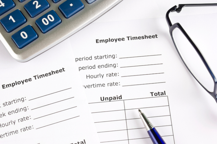 A close-up of employee timesheets with blank fields for period, hourly, and overtime rates. Nearby are glasses, a pen, and a blue calculator. The scene conveys a systematic, organized work environment.